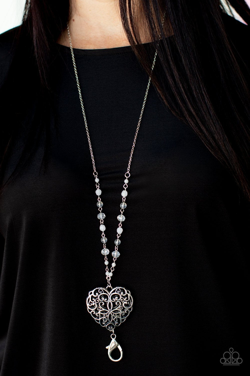 A dainty collection of opaque, glassy, and crystal-like beads give way to an oversized filigree filled heart frame, creating an antiqued locket inspired pendant at the bottom of a lengthened silver chain. A lobster clasp hangs from the bottom of the design to allow a name badge or other item to be attached. Features an adjustable clasp closure. Sold as one individual lanyard. Includes one pair of matching earrings.